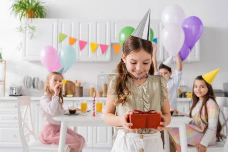 Photo for Pleased girl in party cap holding birthday present near friends on blurred background - Royalty Free Image