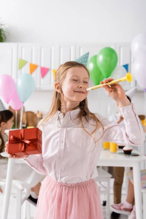 happy preteen girl with closed eyes holding birthday present and party horn near friends on blurred background 