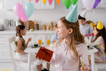 Foto de Feliz niña preadolescente sosteniendo caja de regalo de cumpleaños y cuerno de fiesta cerca de amigos en un fondo borroso - Imagen libre de derechos