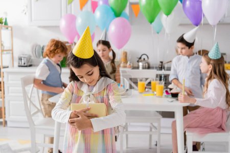 Photo for Happy preteen girl in party cap hugging birthday present near friends on blurred background - Royalty Free Image