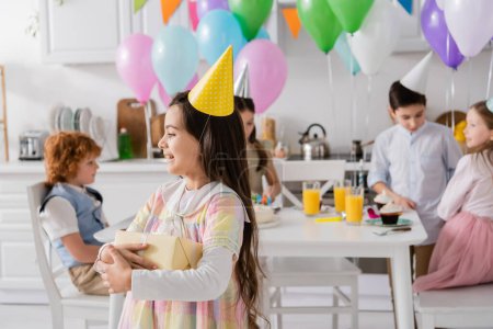 Photo for Cheerful birthday girl in party cap holding gift box near friends on blurred background - Royalty Free Image