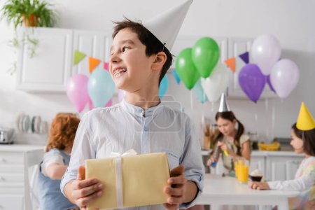 Téléchargez les photos : Joyeux garçon préadolescent dans bretelles tenant cadeau d'anniversaire près d'amis sur fond flou - en image libre de droit