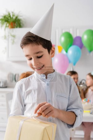 Photo for Smiling preteen boy in braces holding birthday present near friends on blurred background - Royalty Free Image