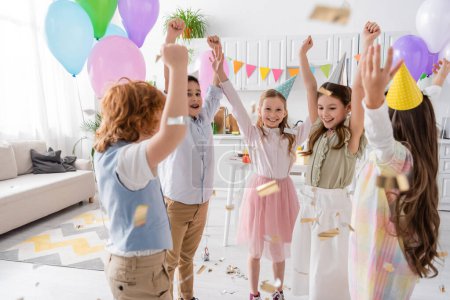 Fröhliche Kinder in Partymützen tanzen bei Geburtstagsfeier zu Hause unter fallendem Konfetti 