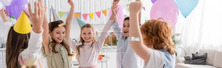 fröhliche Kinder in Partymützen tanzen unter fallendem Konfetti während der Geburtstagsfeier zu Hause, Banner 