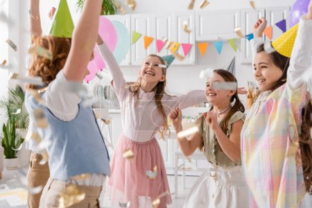 Gruppe aufgeregter Kinder in Partymützen tanzt bei Geburtstagsfeier zu Hause unter fallendem Konfetti 