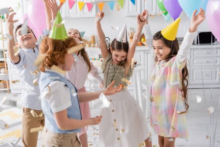 group of joyful kids in party caps dancing under falling confetti during birthday celebration at home 