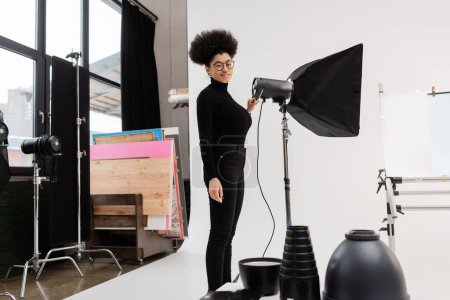 gerente de contenido afroamericano en gafas y ropa negra sonriendo cerca de reflector en estudio fotográfico