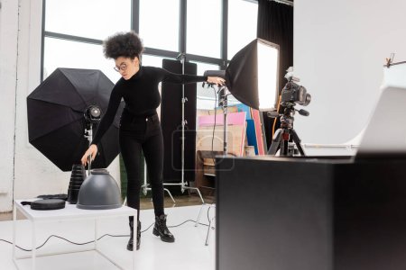 Foto de Productor de contenido afroamericano en ropa negra alcanzando lámparas cerca de reflector softbox y cámara digital en estudio fotográfico moderno - Imagen libre de derechos