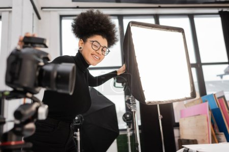 Téléchargez les photos : Joyeux fabricant de contenu afro-américain souriant à la caméra près de projecteur et appareil photo numérique flou dans le studio photo - en image libre de droit