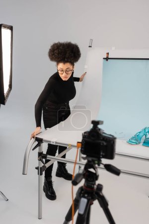 african american content maker adjusting shooting table with trendy sandals near blurred digital camera in photo studio