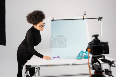 productor de contenido afroamericano mirando gafas de sol y sandalias con estilo en la mesa de tiro cerca de la cámara digital borrosa en el estudio de fotos