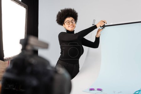 happy african american content producer in eyeglasses installing background sheet in photo studio on blurred foreground