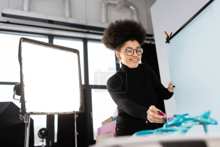 cheerful african american content maker in eyeglasses looking at camera near reflector and shooting table in photo studio