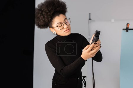 Foto de Elegante gerente de contenido afroamericano en cuello alto negro mirando el medidor de exposición en el estudio de fotos - Imagen libre de derechos