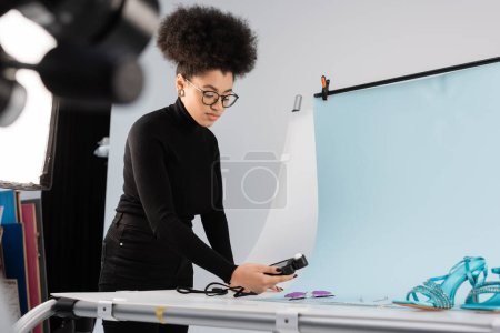 African american content maker using light meter near trendy sunglasses and sandals on shooting table in photo studio