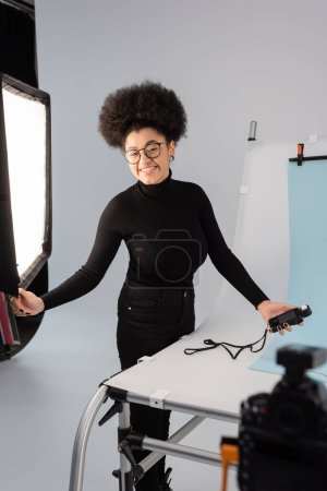 joyful african american content producer with exposure meter looking at camera near spotlight and shooting table in photo studio