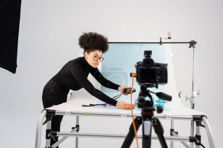 Afrikanisch-amerikanischer Content Maker schaut auf verschwommene Digitalkamera, während er Belichtungsmesser in der Nähe des Aufnahmetisches in einem modernen Fotostudio hält