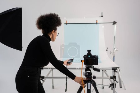 Afrikanischer Produzent von Inhalten hält Belichtungsmesser in der Nähe von Digitalkamera und Aufnahmetisch in modernem Studio