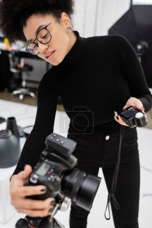 african american content maker in eyeglasses holding exposure meter and adjusting blurred digital camera in photo studio