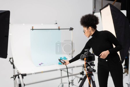 Photo for African american content manager holding exposure meter near sandals and sunglasses on shooting table in photo studio - Royalty Free Image