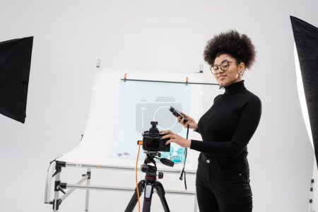 glücklicher und stilvoller afrikanisch-amerikanischer Content Maker mit Belichtungsmesser in der Nähe von Digitalkamera und Aufnahmetisch im modernen Studio