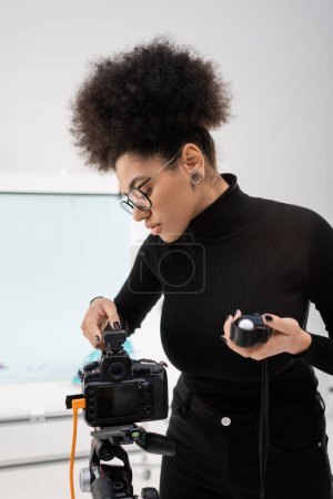 Téléchargez les photos : Élégant gestionnaire de contenu afro-américain tenant compteur d'exposition et réglage de l'appareil photo numérique dans le studio photo - en image libre de droit