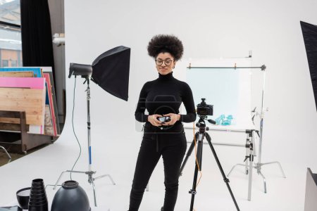 producteur de contenu afro-américain souriant debout avec compteur d'exposition près de l'appareil photo numérique et de l'équipement d'éclairage dans un studio photo moderne