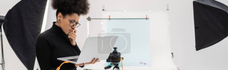Photo for Serious african american content producer looking at laptop while thinking near reflectors and digital camera in photo studio, banner - Royalty Free Image