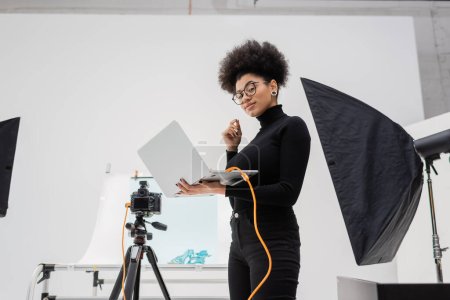 fabricante de contenido afroamericano con portátil sonriendo cerca de softbox y cámara digital en estudio de fotos contemporáneo
