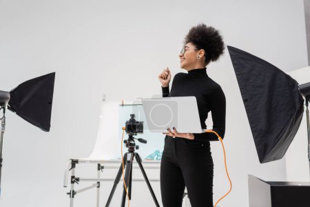 Foto de Fabricante de contenido afroamericano alegre con portátil mirando hacia otro lado cerca de softboxes y cámara digital en el estudio de fotos moderno - Imagen libre de derechos