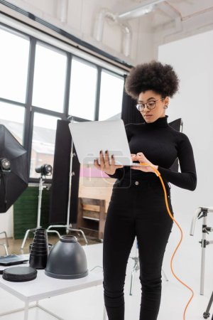 Photo for African american content manager in eyeglasses using laptop while standing near lighting equipment in photo studio - Royalty Free Image
