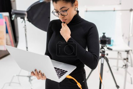 nachdenkliche afrikanisch-amerikanische Content Maker in Brille Blick auf Laptop in der Nähe verschwommene Digitalkamera in Fotostudio