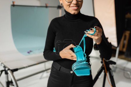 Photo for Cropped view of smiling african american content maker in black turtleneck holding trendy sandal in blurred photo studio - Royalty Free Image