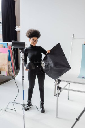 in voller Länge lächelnder afrikanisch-amerikanischer Content Maker in schwarzer Kleidung mit Softbox in der Nähe von Scheinwerfern im Fotostudio
