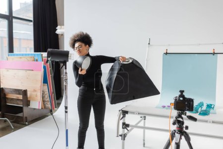 producteur de contenu afro-américain assemblant des équipements d'éclairage et tenant réflecteur dans un studio moderne