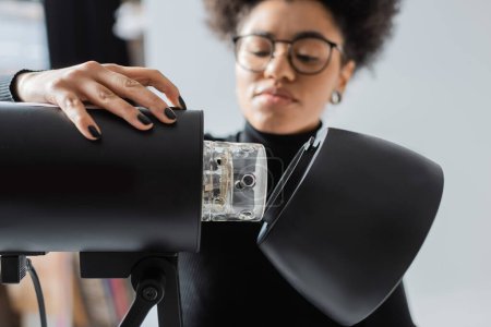 Téléchargez les photos : Flou afro-américain producteur de contenu dans les lunettes montage lampe stroboscopique en studio photo - en image libre de droit