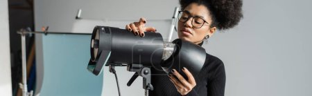 fabricant de contenu afro-américain élégant dans les lunettes de travail avec un équipement d'éclairage professionnel dans le studio photo, bannière
