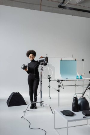 longitud completa de fabricante de contenido afroamericano feliz cerca de la lámpara estroboscópica y la mesa de tiro en el estudio de fotos contemporáneo