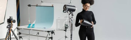 african american content manager assembling strobe lamp near digital camera and shooting table in photo studio, banner