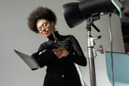 african american content producer in eyeglasses holding part of strobe lamp in photo studio