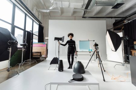 in voller Länge überglücklicher afrikanisch-amerikanischer Content Manager in schwarzer Kleidung, der neben Stroboskoplampe im modernen Studio steht