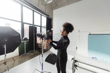 Téléchargez les photos : Vue latérale du créateur de contenu afro-américain ajustant la lampe stroboscopique tout en travaillant dans un studio photo contemporain - en image libre de droit
