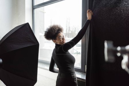cheerful african american content producer pulling curtain near window in photo studio and smiling at camera