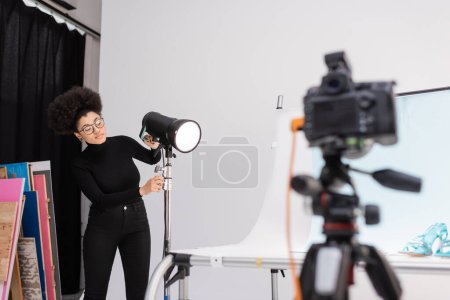 fabricante de contenido afroamericano ajustando el reflector cerca de la mesa de tiro y la cámara digital borrosa en el estudio de fotos