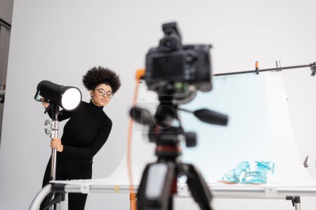 afrikanisch-amerikanischer Content Manager justiert Blitzlampe in der Nähe verschwommener Digitalkamera und Aufnahmetisch mit stylischem Schuhwerk im Fotostudio