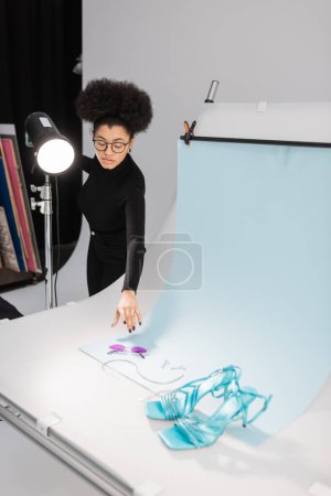 Téléchargez les photos : Afro-américain producteur de contenu près de lampe stroboscopique et table de tir avec des lunettes de soleil élégantes et des sandales dans le studio photo - en image libre de droit