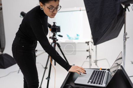 african american content producer in eyeglasses using laptop near reflector and blurred digital camera in photo studio