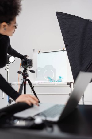 Téléchargez les photos : Afro-américain fabricant de contenu réglage appareil photo numérique près de la table de tir avec des chaussures à la mode et un ordinateur portable flou dans le studio photo - en image libre de droit