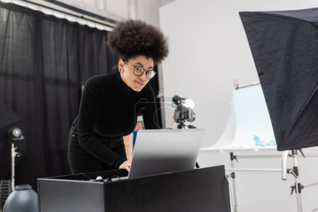 Photo for Smiling african american content manager in eyeglasses and black turtleneck looking at camera near laptop in photo studio - Royalty Free Image
